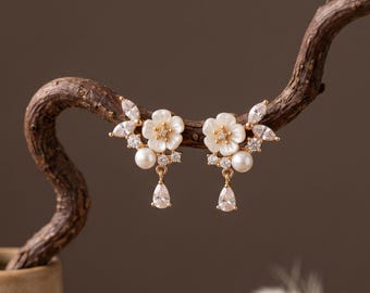 Può includere: Un paio di orecchini color oro con un motivo floreale bianco centrale, impreziositi da piccole perle rotonde, cristalli trasparenti taglio marquise e una singola goccia di cristallo pendente a forma di pera. Il design è simmetrico e raffinato.