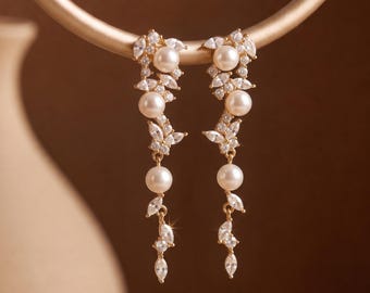 Puede incluir: Un par de pendientes colgantes en tono dorado con una disposición vertical de perlas blancas redondas y cristales transparentes de talla marquesa. El diseño presenta una estructura similar a una rama con perlas y piedras brillantes.