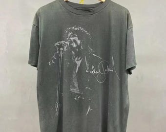 May include: A gray t-shirt featuring a black and white image of a singer performing with a microphone. The singer's signature is printed in white. The shirt is hanging on a wooden hanger.