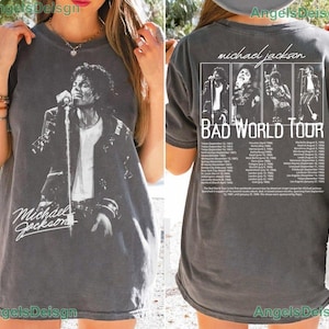 May include: A charcoal gray t-shirt featuring a black and white graphic of Michael Jackson on the front with his name in script. The back displays four black and white photos of the artist, the text "michael jackson BAD WORLD TOUR", and a list of tour dates.