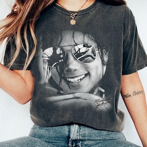 May include: A charcoal grey short-sleeve t-shirt featuring a black and white photographic print of Michael Jackson wearing sunglasses. The graphic includes the name Michael Jackson in a cursive script font located in the lower right corner.