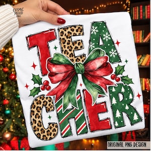 May include: A white t-shirt featuring the word TEA in red and leopard print, and CHER in leopard print, red, and green with white Christmas trees. A large red and green bow sits in the centre, surrounded by holly leaves and red berries.