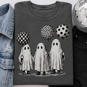 May include: A charcoal gray t-shirt featuring a graphic illustration of three white ghosts standing together. Each ghost holds a balloon with a different pattern: one checkered, one leopard print, and one polka dot. The design is centered on the shirt.