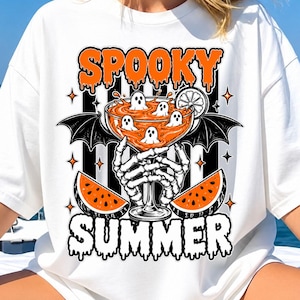 Op de afbeelding: Een wit t-shirt met een grafische print van een skeletthand die een cocktailglas vasthoudt gevuld met oranje vloeistof en kleine spookjes. Het ontwerp bevat twee watermeloenpartjes, vleermuisvleugels en de tekst SPOOKY SUMMER in een druipend lettertype.