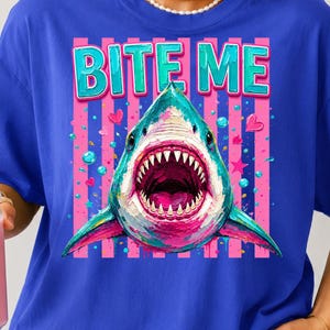 May include: A royal blue t-shirt featuring a graphic of a shark with its mouth open, displaying sharp teeth. The text BITE ME is printed in teal above the shark. The background of the graphic consists of vertical pink stripes, small hearts, and stars.