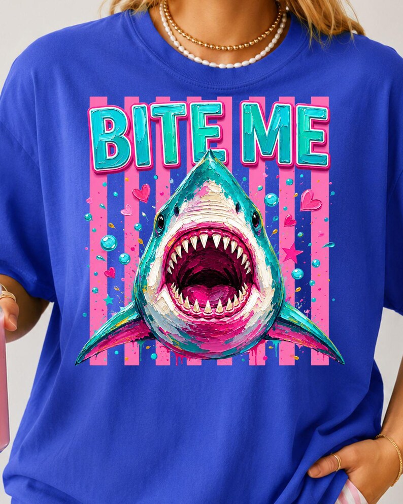 May include: A royal blue t-shirt featuring a graphic of a shark with its mouth open, displaying sharp teeth. The text BITE ME is printed in teal above the shark. The background of the graphic consists of vertical pink stripes, small hearts, and stars.