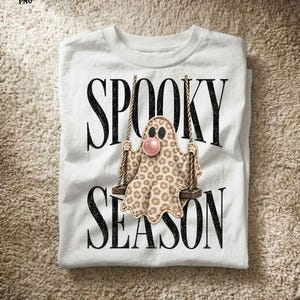 May include: A white t-shirt featuring the black text SPOOKY SEASON. Centered is a graphic of a ghost with a tan leopard print pattern sitting on a wooden swing, blowing a pink bubble. The ghost has two black eyes and a stitched outline effect.