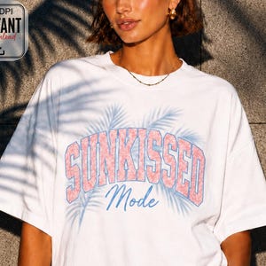 以下が含まれることがあります： ピンクと青のデザインで「Sunkissed Mode」の文字が入った白いTシャツ。 ゆったりとしたフィット感と半袖です。 デザインにはヤシの葉が含まれています。 画像には「300 DPI Instant Download」グラフィックも含まれています。