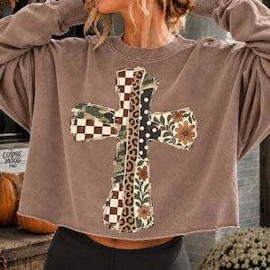 May include: A brown long-sleeve sweatshirt featuring a large cross graphic composed of vertical panels with checkered, camouflage, leopard print, polka dot, and floral patterns. The text COSMIC MOOD PNG is printed in black on the left side.