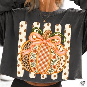 May include: A charcoal gray crewneck sweatshirt featuring a large graphic of a pumpkin with a light pink bow. The pumpkin has sections with leopard print, orange gingham, and floral patterns. Behind the pumpkin are vertical cream stripes with brown polka dots.