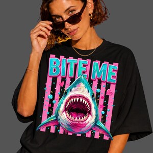 May include: A black t-shirt featuring a graphic print of a shark with an open mouth, surrounded by pink and blue bubbles, stars, and hearts. Above the shark, the text BITE ME is printed in large, bold, light blue letters with a pink outline.