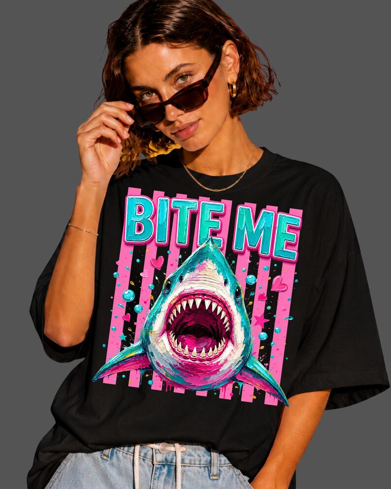 May include: A black t-shirt featuring a graphic print of a shark with an open mouth, surrounded by pink and blue bubbles, stars, and hearts. Above the shark, the text BITE ME is printed in large, bold, light blue letters with a pink outline.