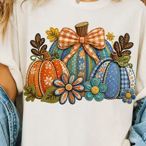 Peut inclure: Un t-shirt couleur crème arborant un motif de trois citrouilles aux motifs variés, incluant des imprimés floraux et vichy. Les citrouilles sont décorées d'un grand nœud vichy orange et blanc, de feuilles vertes et de deux fleurs bleu et orange.
