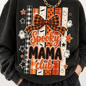 Può includere: Una felpa nera a girocollo con una stampa grafica di strisce verticali in motivi arancioni, neri e bianchi. Il design include un grande fiocco a quadri arancione e nero, piccoli fantasmi bianchi e stelle nere, con il testo Spooky MAMA club.