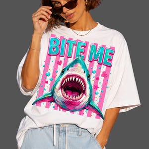 May include: A white short-sleeve t-shirt featuring a graphic print of a shark with an open mouth. The design includes the text "BITE ME" in blue and pink letters, set against a background of vertical pink stripes, small hearts, and stars.