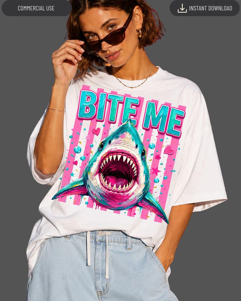 May include: A white short-sleeve t-shirt featuring a graphic print of a shark with an open mouth. The design includes the text "BITE ME" in blue and pink letters, set against a background of vertical pink stripes, small hearts, and stars.