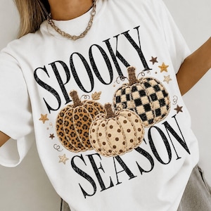 May include: A white t-shirt featuring the black text SPOOKY SEASON and three pumpkins. One pumpkin has a brown leopard print, one has a black and white checkered pattern, and one has a tan polka dot design, accented with small gold stars.