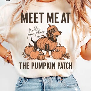 May include: A cream-colored t-shirt featuring a graphic of a dachshund wearing a pumpkin-shaped hat and sweater, surrounded by pumpkins. The text reads "MEET ME AT", "hello pumpkin", and "THE PUMPKIN PATCH" in dark brown lettering.