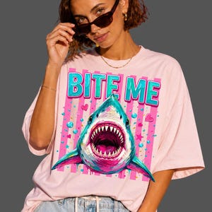 May include: A light pink t-shirt featuring a graphic print of a shark with an open mouth and sharp teeth. Above the shark, the text BITE ME is printed in a bold, blue, three-dimensional font. The background of the graphic includes pink stripes.