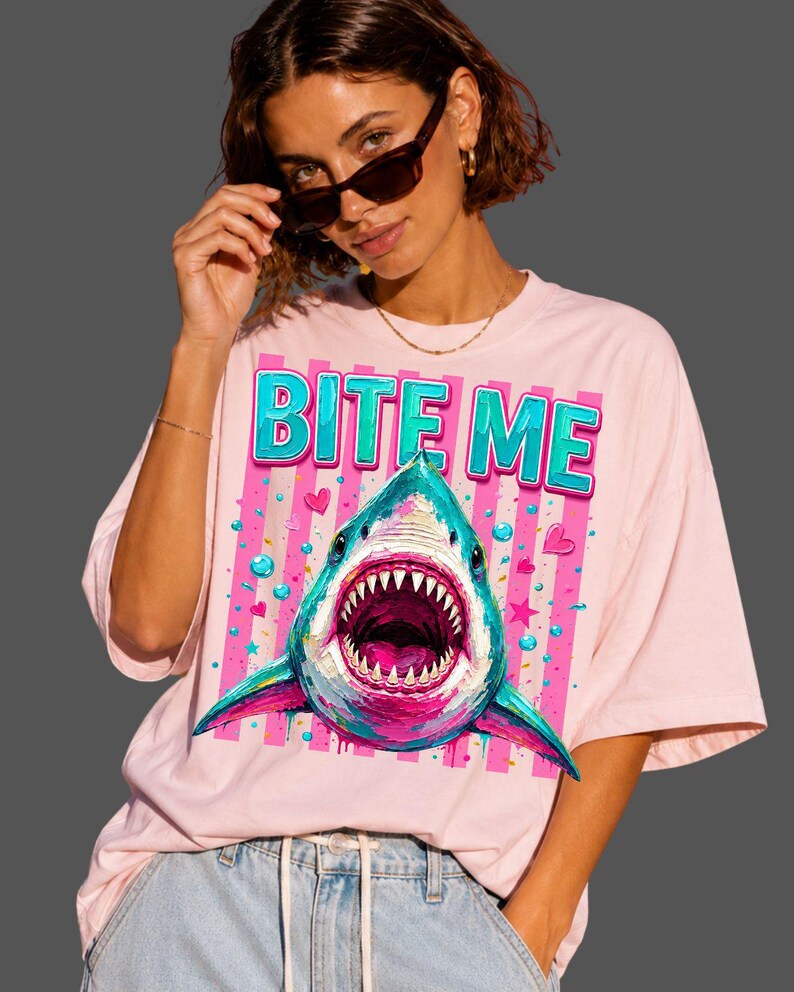 May include: A light pink t-shirt featuring a graphic print of a shark with an open mouth and sharp teeth. Above the shark, the text BITE ME is printed in a bold, blue, three-dimensional font. The background of the graphic includes pink stripes.