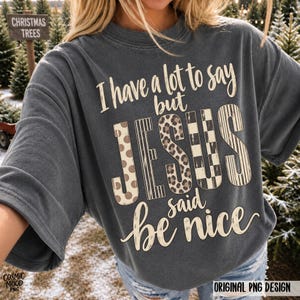 May include: A charcoal gray short-sleeve t-shirt featuring the text I have a lot to say but JESUS said be nice. The word JESUS is printed in large letters with varying patterns including polka dots, vertical stripes, leopard print, and checkers.