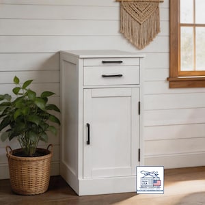 May include: A white wooden bedside cabinet with a drawer and a door. The cabinet has black handles and is placed next to a potted plant in a woven basket. A macrame wall hanging and a window are visible in the background.