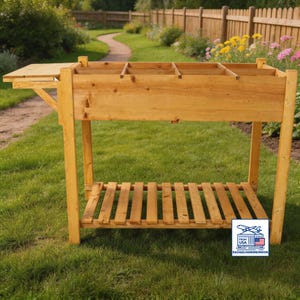 May include: A wooden potting bench with a natural finish. It features a rectangular top with divided sections, a pull-out side table, and a slatted shelf. The bench stands on four sturdy legs and is set on a grassy lawn.