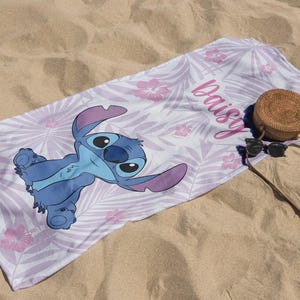 May include: A beach towel featuring a blue cartoon character with large ears, lying on a white and lavender floral patterned background. The name "Daisy" is written in pink script. A round woven bag and sunglasses rest on the sand.