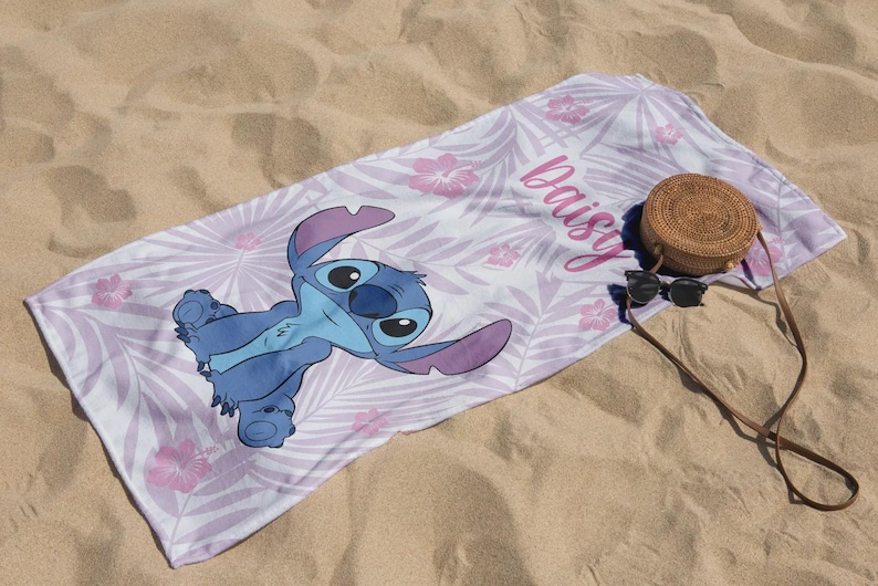 May include: A beach towel featuring a blue cartoon character with large ears, lying on a white and lavender floral patterned background. The name "Daisy" is written in pink script. A round woven bag and sunglasses rest on the sand.