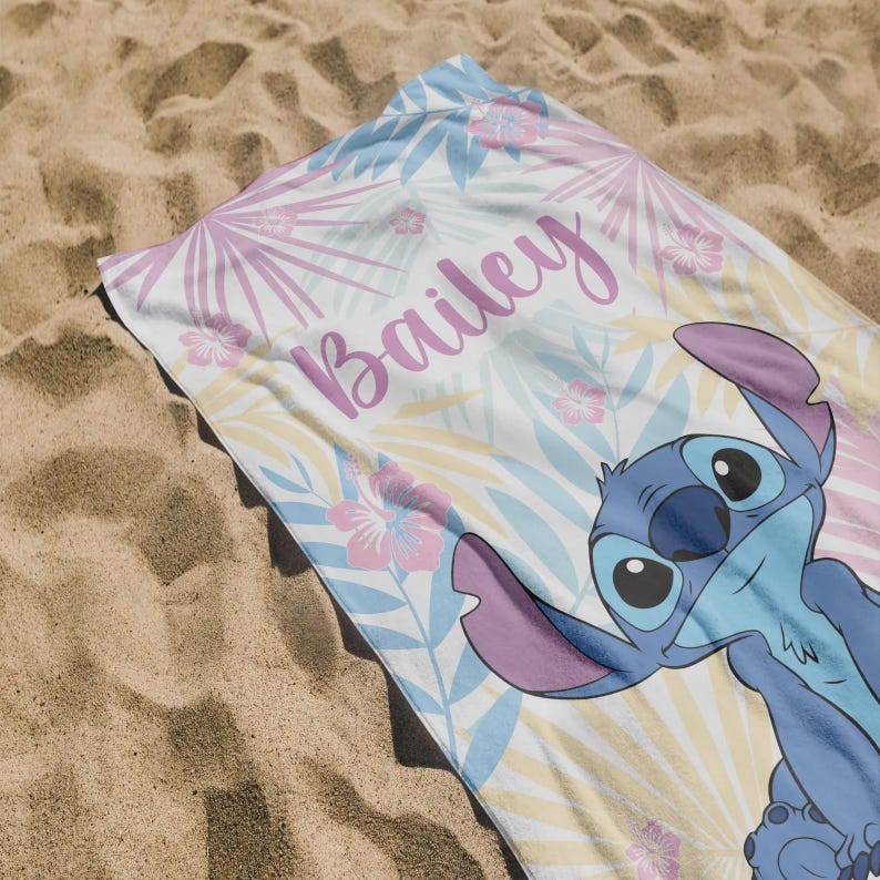 May include: A beach towel featuring a cartoon character in blue with large ears, set against a tropical floral pattern in pastel colors. The name "Bailey" is written in cursive. The towel is on a sandy surface.