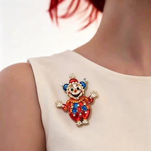 May include: A vibrant clown brooch in red, blue, and gold. The brooch depicts a smiling clown with a red hat, blue ears, and a gem. The clown's outfit is red and blue with a diamond pattern. A fun and decorative accessory.