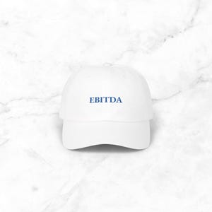 May include: A white baseball cap with a curved peak. The cap displays the word "EBITDA" in blue block letters. The hat is made of a soft material and is suitable for casual wear.