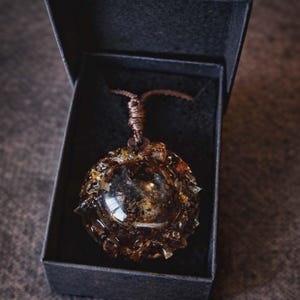 Może przedstawiać: Ręcznie robiony naszyjnik z wisiorkiem o unikalnym, kulistym kształcie. Wisiorek ma centralny, przezroczysty, sferyczny element otoczony teksturowanym materiałem w kolorze bursztynu. Naszyjnik jest prezentowany w czarnym pudełku.