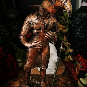 May include: A bronze-colored sculpture of a figure with a bird-like head, holding a sword. The figure stands on a textured base with a circular emblem. The sculpture has a detailed, aged appearance, with a second bird head behind the figure.
