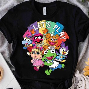 Peut inclure: Un t-shirt noir à manches courtes avec un graphique coloré des personnages de Muppet Babies, dont Kermit, Piggy, Fozzie, Gonzo, Animal et Summer. Le texte LET'S PLAY est imprimé en blanc au-dessus des personnages sur un fond arc-en-ciel.