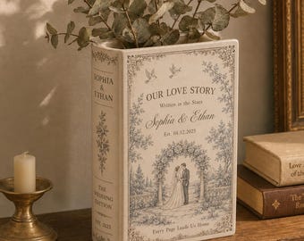 Puede incluir: Un jarrón de cerámica rectangular con forma de libro, acabado en color crema e ilustraciones azules. El frente presenta el texto "Our Love Story", "Written in the Stars", "Sophia & Ethan", "Est. 04.12.2025" y "Every Page Leads Us Home".