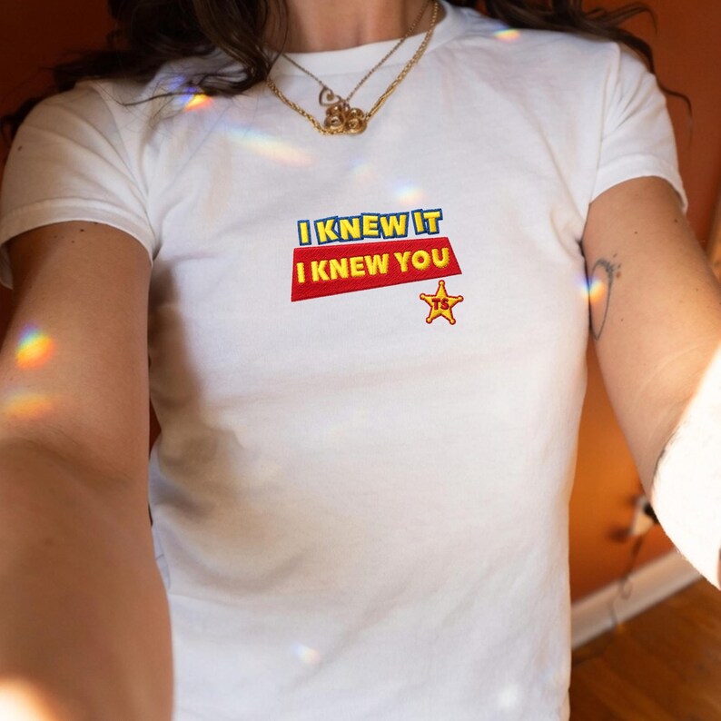 K&ouml;nnte beinhalten: Ein wei&szlig;es kurz&auml;rmeliges T-Shirt mit einer gestickten Grafik auf der Brust. Das Design zeigt den blauen Schriftzug I KNEW IT &uuml;ber dem roten Schriftzug I KNEW YOU sowie einen gelben Sheriffstern mit den Buchstaben TS darunter.