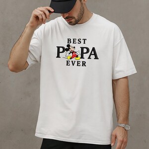 Puede incluir: Camiseta blanca con el texto negro "BEST PA EVER", con un dibujo animado de Mickey Mouse. La camiseta es de estilo casual y de gran tama&ntilde;o.