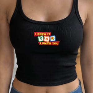 May include: A black ribbed tank top featuring a central graphic with the text I KNEW IT and I KNEW YOU in yellow letters on red rectangular backgrounds, surrounding three colorful square blocks displaying the letters T, S, and a red star icon.