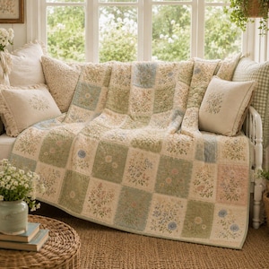 May include: A patchwork quilt featuring a grid of squares with various floral prints in muted shades of green, blue, and pink. The quilt is draped over a white wooden frame, accompanied by two matching square throw pillows with floral embroidery.