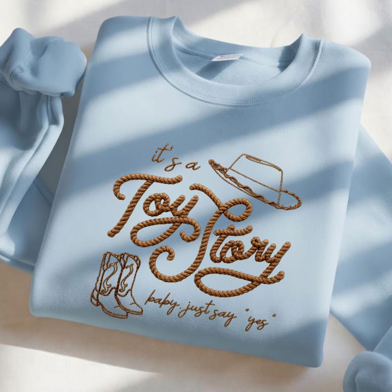 Peut inclure: Un sweat-shirt bleu clair &agrave; col rond avec une broderie marron en forme de corde formant l'inscription "it's a Toy Story". Le motif comprend un chapeau de cowboy brod&eacute; au-dessus du texte et des bottes de cowboy en dessous, avec "baby just say yes".