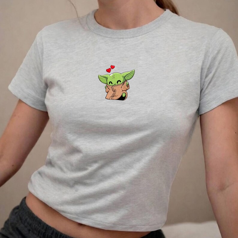 May include: A light grey crop top with a cartoon illustration of a green alien character wearing a tan robe. Two small red hearts float above the character's head. The top has short sleeves.