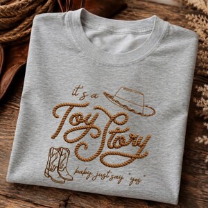 Peut inclure: Un t-shirt gris clair &agrave; col rond avec un texte brod&eacute; marron indiquant "it's a Toy Story" et "baby just say yes". Le motif comprend un chapeau de cowboy et une paire de bottes de cowboy brod&eacute;s en marron, avec une texture de corde.