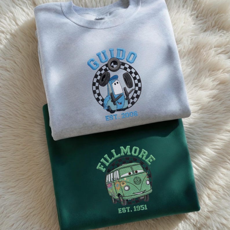 May include: Two sweatshirts, one light gray and one dark green, are stacked on a fluffy white surface. The light gray sweatshirt features a blue embroidered design of a car named "Guido" with the text "EST. 2006". The dark green sweatshirt has a green bus design with the text "FILLMORE EST. 1951".