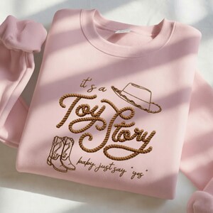 Peut inclure: Un sweat-shirt &agrave; col rond rose clair avec une broderie marron formant le texte "it's a Toy Story" dans une police style corde. Le motif comprend un chapeau de cowboy et une paire de bottes de cowboy brod&eacute;s, avec le texte "baby, just say 'yes'" en dessous.