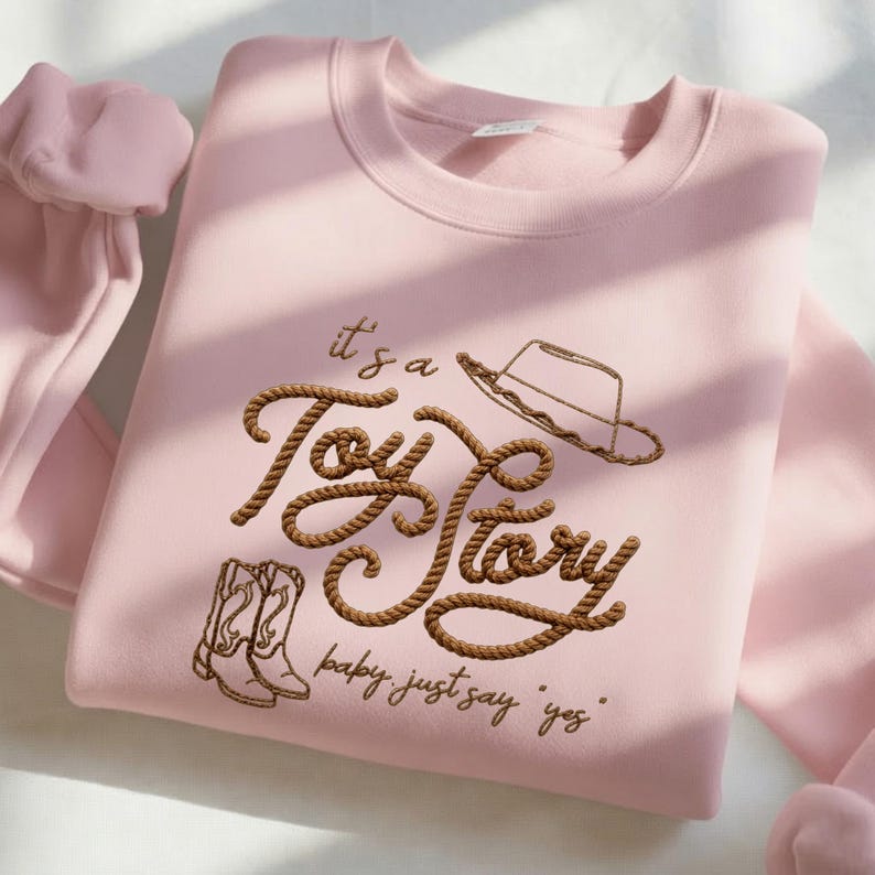 Peut inclure: Un sweat-shirt &agrave; col rond rose clair avec une broderie marron formant le texte "it's a Toy Story" dans une police style corde. Le motif comprend un chapeau de cowboy et une paire de bottes de cowboy brod&eacute;s, avec le texte "baby, just say 'yes'" en dessous.