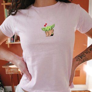May include: A pale pink t-shirt with a cartoon character embroidered on the chest. The character has green skin, large ears, and a tan robe. Two small red hearts float above the character's head. The t-shirt has short sleeves.