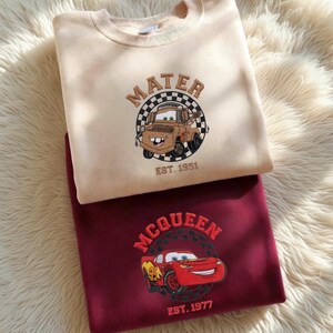 May include: Two sweatshirts, one beige and one burgundy, are displayed. The beige sweatshirt features an embroidered image of Mater from the movie Cars, with the text "MATER" and "EST. 1951." The burgundy sweatshirt has an embroidered image of Lightning McQueen, with the text "MCQUEEN" and "EST. 1977."