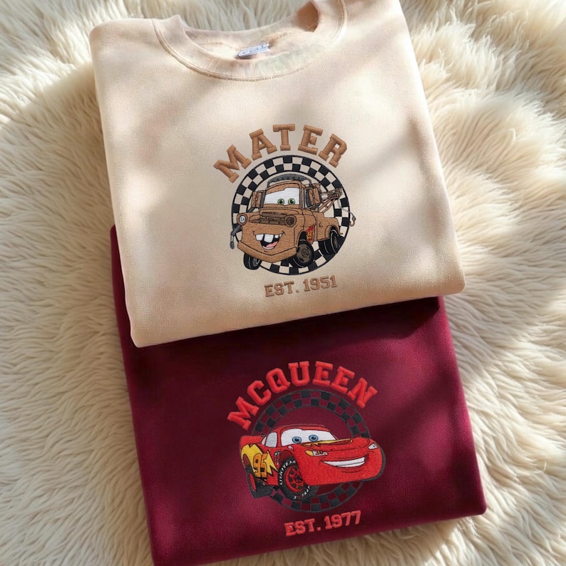 May include: Two sweatshirts, one beige and one burgundy, are displayed. The beige sweatshirt features an embroidered image of Mater from the movie Cars, with the text "MATER" and "EST. 1951." The burgundy sweatshirt has an embroidered image of Lightning McQueen, with the text "MCQUEEN" and "EST. 1977."