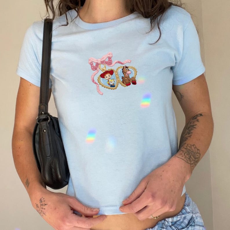 May include: Light blue t-shirt with a crew neck and short sleeves. The shirt features an embroidered design of two heart-shaped frames with cartoon characters and a pink bow. The shirt is made of a soft material.